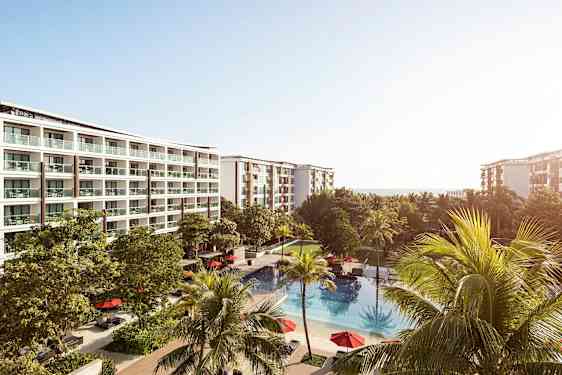 Et hotellkompleks med flere etasjer og balkonger, omgitt av grønne trær. Foran bygningene ligger et utendørsbasseng med røde parasoller. (AI-generert tekst)