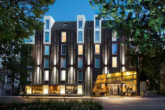 Eksteriør av et moderne hotell med glassfasade og belyste vinduer omgitt av trær. (AI-generert tekst)