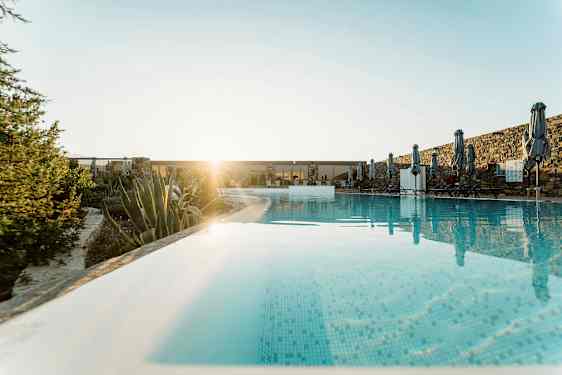 Et svømmebasseng med parasoller og solsenger, omgitt av planter og steinmur, med solen i bakgrunnen. (AI-generert tekst)