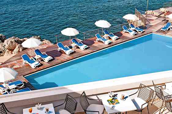 Et basseng med solsenger og parasoller ved en treterrasse ved havet. Forgrunnen viser et bord med stoler og drikker. (AI-generert tekst)