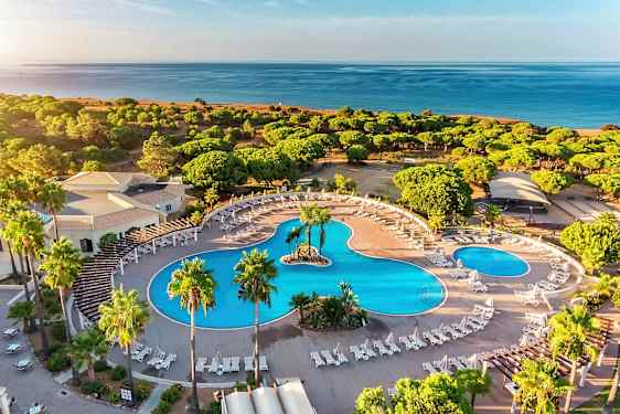Flyfoto av et hotellområde med store bassenger, solsenger og omkringliggende grøntområde nær stranden. (AI-generert tekst)