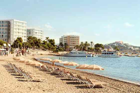 Strand med solsenger og parasoller, hotellbygninger i bakgrunnen og båter på vannet. (AI-generert tekst)