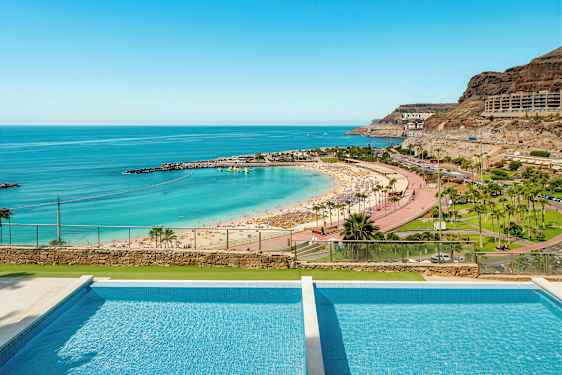 Bilde av en strand med parasoller, turkis vann og et basseng i forgrunnen. (AI-generert tekst)