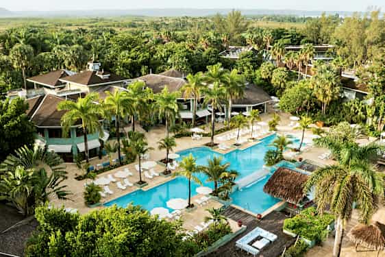 Flyfoto av et hotellområde med et stort basseng omgitt av solsenger og parasoller, samt frodige trær og vegetasjon. (AI-generert tekst)