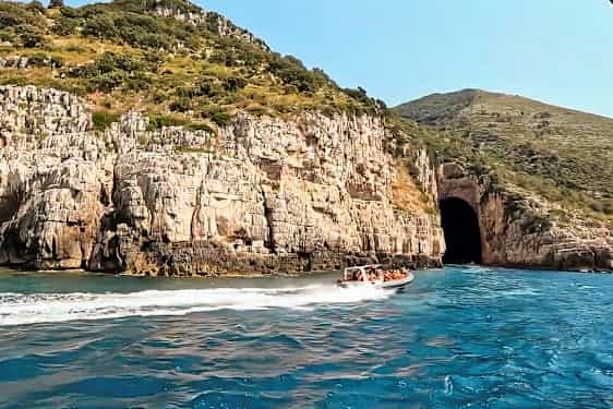 En motorbåt med folk kjører på blått vann nær en klippe med en stor hulåpning. (AI-generert tekst)