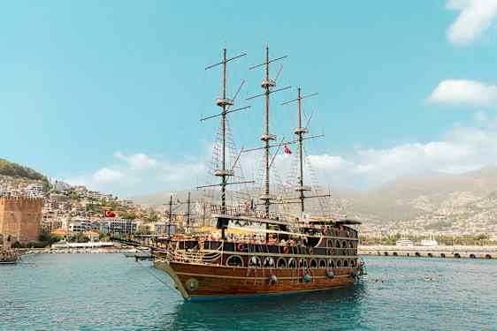 Et stort seilskip ligger ved havnen, med en by og grønne åser i bakgrunnen. (AI-generert tekst)