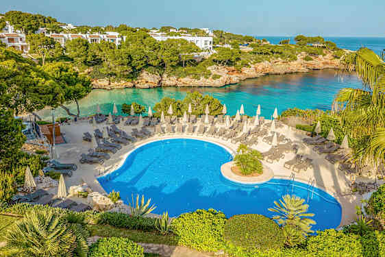 Et utendørs svømmebasseng med solsenger og parasoller, omgitt av grønne områder og med utsikt over havet. (AI-generert tekst)