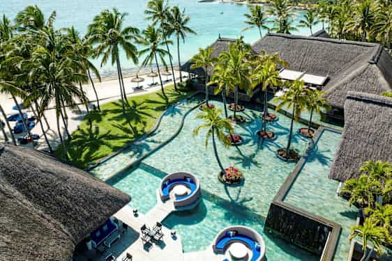 Flere hus med halmtak ved et basseng med palmer og sandstrand i bakgrunnen. (AI-generert tekst)