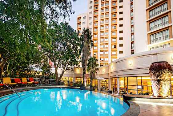 Et opplyst utendørsbasseng omgitt av grønne planter og en bygning i bakgrunnen. (AI-generert tekst)