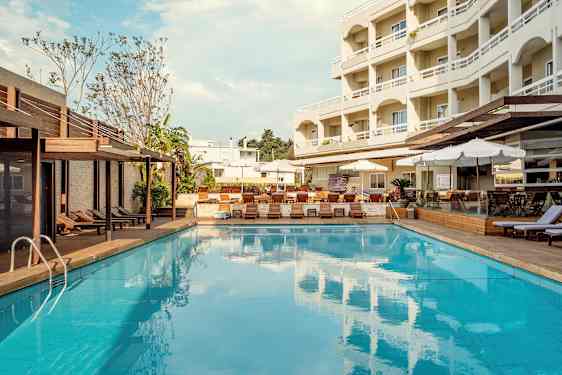 Et utendørsbasseng med solsenger og parasoller på siden og et fleretasjes hotell i bakgrunnen. (AI-generert tekst)