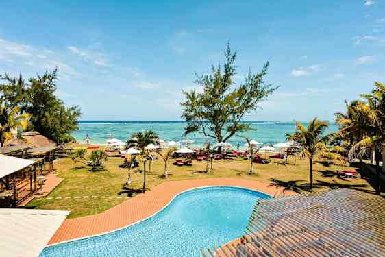 Et basseng med turkist vann foran en plen med trær, solsenger og parasoller langs en strand. (AI-generert tekst)