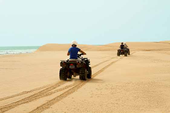 To personer kjører firehjulinger over en sandstrand med havet i bakgrunnen. (AI-generert tekst)