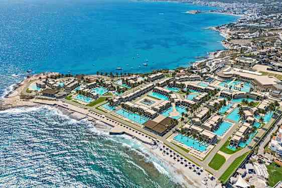 Et dronebilde av en ferieanlegg ved kysten med bassenger, bygninger og stranden ved siden av havet. (AI-generert tekst)