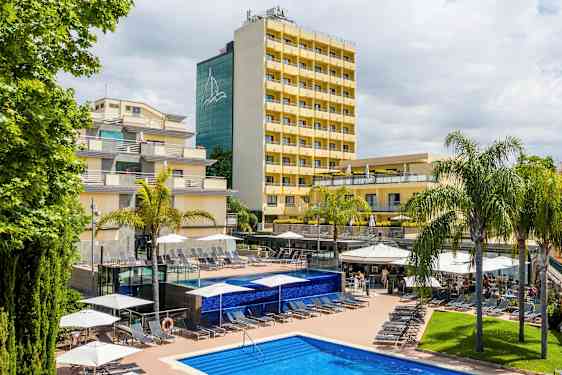 Bilde av et hotell med gule fasader, omgitt av palmer og et basseng med solsenger på en solterrasse. (AI-generert tekst)