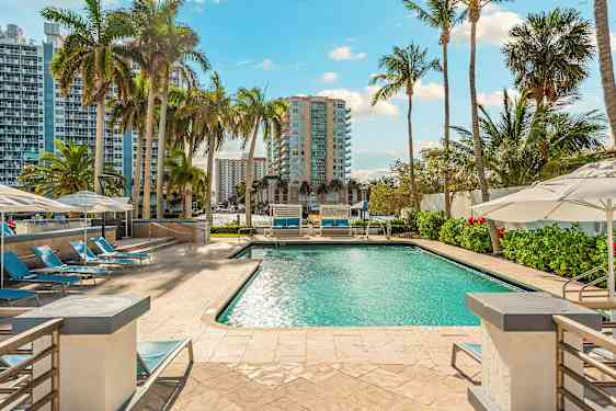 Et svømmebasseng omgitt av solsenger og parasoller, med høye bygninger og palmer i bakgrunnen. (AI-generert tekst)