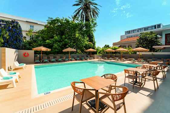 Et utendørsbasseng med solsenger og parasoller på siden samt bord og stoler i forgrunnen. (AI-generert tekst)