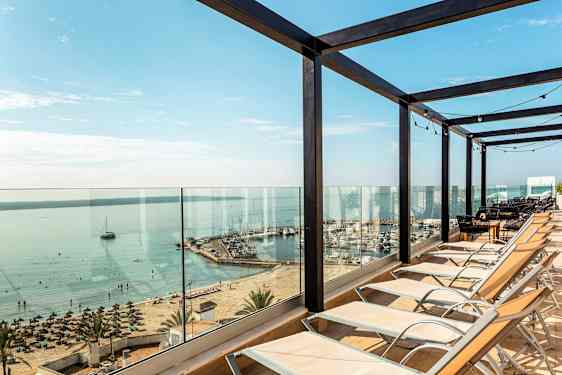 Solstoler satt opp foran glassrekkverk med utsikt over strand og havn. (AI-generert tekst)