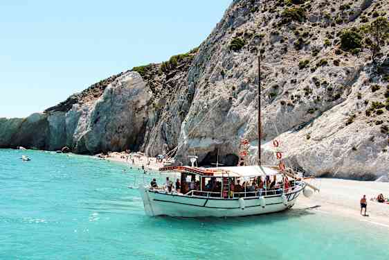 En båt ligger fortøyd ved en strand med klipper og klart turkisblått vann. (AI-generert tekst)