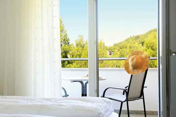 Et hotellrom med utsikt til en balkong hvor det står en stol med en hatt og et bord, samt frodige trær i bakgrunnen. (AI-generert tekst)