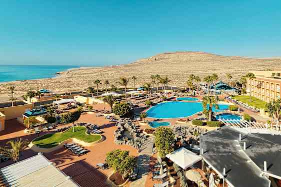 Bildet viser et ferieanlegg ved en kyst med bassengområder, solsenger og palmer. I bakgrunnen sees havet og det omkringliggende tørre landskapet. (AI-generert tekst)