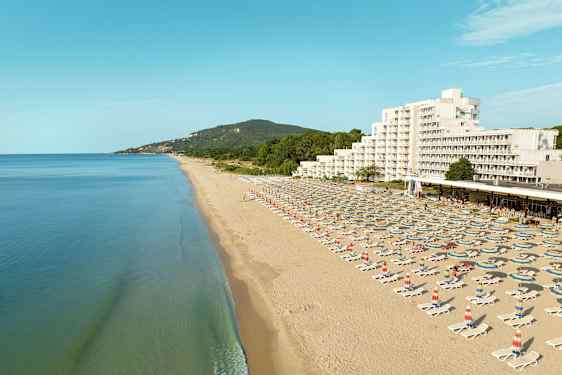 En lang sandstrand med solsenger og parasoller, samt et hotell og grønne åser i bakgrunnen. (AI-generert tekst)