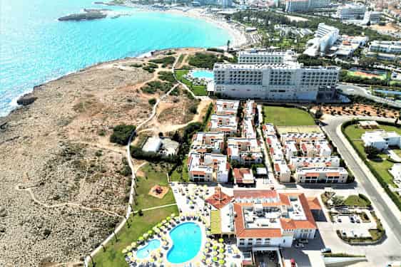 Flybilde av hotellkomplekset med basseng, omkringliggende bygninger og en steinete kyst med utsikt over havet. (AI-generert tekst)