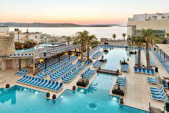 Stort bassengområde med solsenger, palmer og utsikt over havet ved solnedgang. (AI-generert tekst)