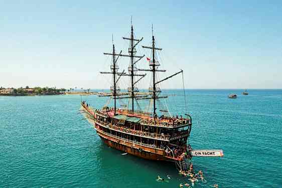 Et stort treskip med seil som ligger i rolig vann, omgitt av svømmere, og i bakgrunnen sees kystlinjen. (AI-generert tekst)