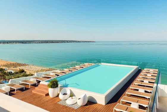 Et basseng på taket med solsenger ved kanten og utsikt over en strand og hav. (AI-generert tekst)