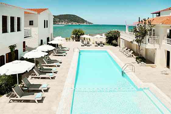 Bilde av et utendørs svømmebasseng med solsenger og parasoller, omgitt av hvite bygninger med utsikt over havet. (AI-generert tekst)
