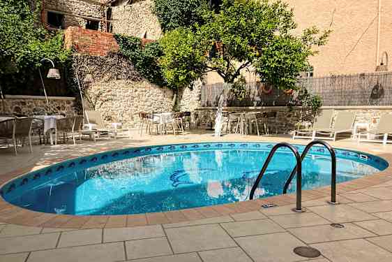 Utendørsbilde med et basseng omgitt av solsenger og bord nær en steinmur og grønne planter. (AI-generert tekst)