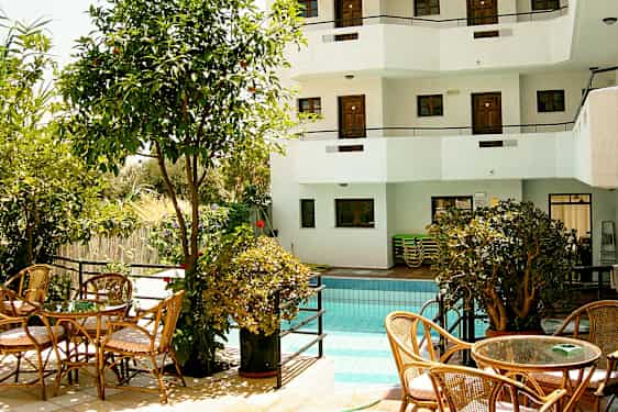 Et utendørs basseng omgitt av vegetasjon og sittemøbler, samt en hvit bygning med balkonger i bakgrunnen. (AI-generert tekst)