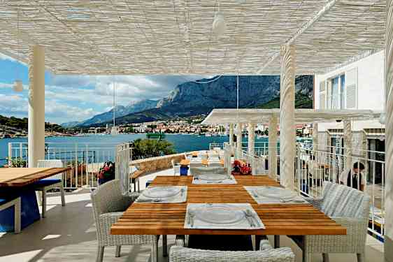 En terrasse med utsikt over fjell og hav, flere bord er dekket for gjester. (AI-generert tekst)