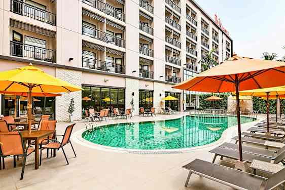 Et hotellbasseng omgitt av solsenger og parasoller, med bygningen i bakgrunnen. (AI-generert tekst)