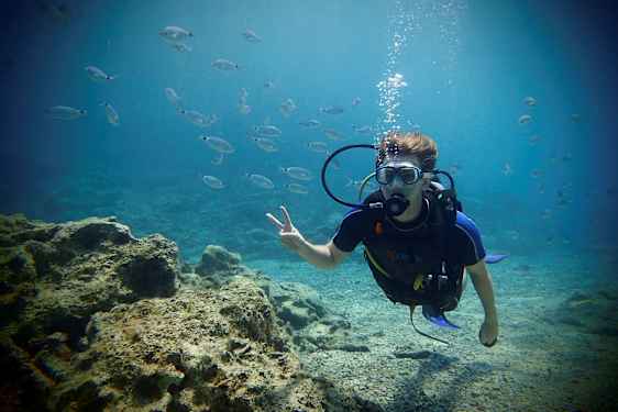 En person i dykedrakt under vann omgitt av små fisker og med en steinet havbunn i bakgrunnen. (AI-generert tekst)