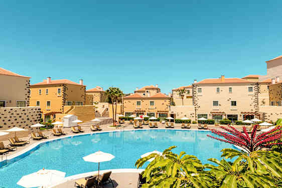 Et basseng omgitt av gule bygninger og solsenger under parasoller. (AI-generert tekst)