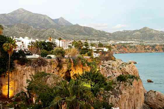 En utsikt over klipper med hus og frodig vegetasjon, med fjell i bakgrunnen og havet til høyre. (AI-generert tekst)