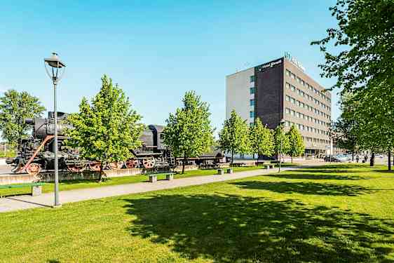 Et gammelt damplokomotiv står utendørs ved siden av et moderne hotellanlegg og frodige trær. (AI-generert tekst)
