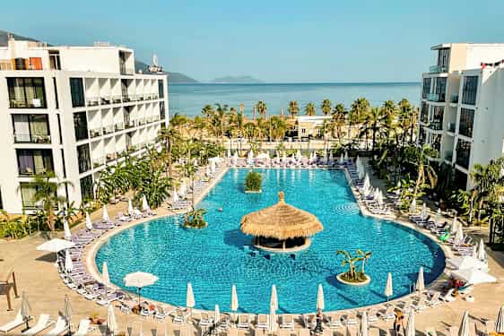 Hotellkompleks med stort basseng omgitt av solsenger og parasoller, med havet i bakgrunnen. (AI-generert tekst)