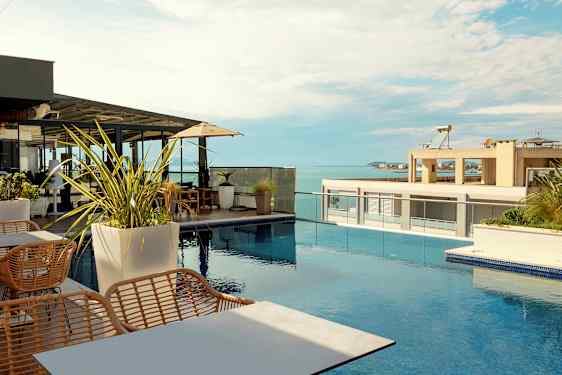 En takterrasse med utsikt over havet, møblert med bord, stoler og parasoller ved et svømmebasseng. (AI-generert tekst)