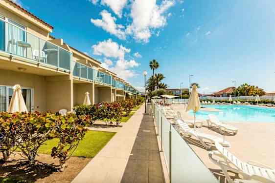 Bilde av et hotellområde med basseng, solsenger og grøntområder. Bygningen har balkonger. (AI-generert tekst)