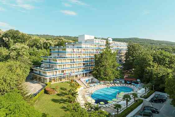 Bilde av et hotell med flere etasjer og balkonger, omgitt av frodig grønt. Et basseng med solsenger ligger i forgrunnen. (AI-generert tekst)
