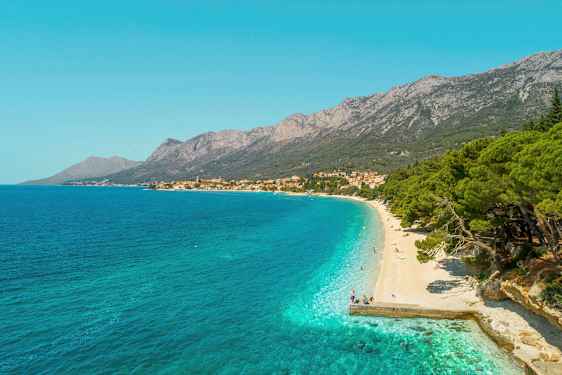 Utsikt over en rullesteinstrand med turkist vann og frodige trær. Fjell og en landsby vises i bakgrunnen. (AI-generert tekst)
