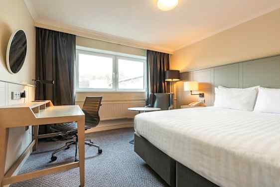 Bilde av et hotellrom med en seng, skrivebord og stol, og en lenestol ved siden av et vindu. (AI-generert tekst)