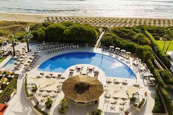 Utsikt over et bassengområde med solsenger og parasoller, omgitt av grønne områder og nær stranden. (AI-generert tekst)