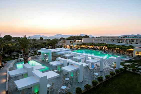 Moderne hotellområde med basseng og kveldsbelysning, omgitt av grøntområder og palmer med fjell i bakgrunnen. (AI-generert tekst)