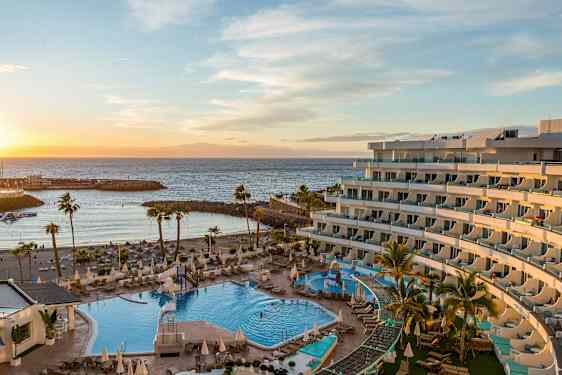 Hotellområde ved sjøen med utendørsbasseng og solsenger, omgitt av palmer, med en strand og bølgebryter i bakgrunnen ved solnedgang. (AI-generert tekst)