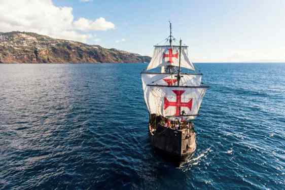 Et seilskip med store hvite seil dekorert med røde kors navigerer over et blått hav nær en kupert kyst. (AI-generert tekst)