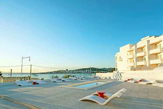 Utendørs bassengområde med moderne solsenger og utsikt over sjøen og hvite bygninger i bakgrunnen. (AI-generert tekst)