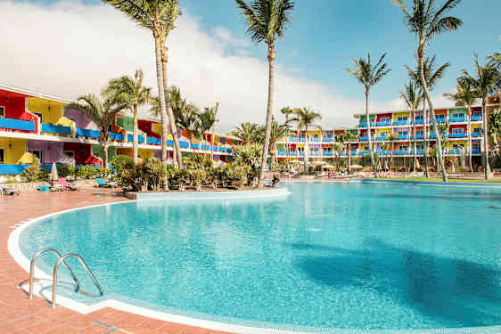 Et stort basseng omgitt av palmer, med fargerike hotellbygninger i bakgrunnen. (AI-generert tekst)